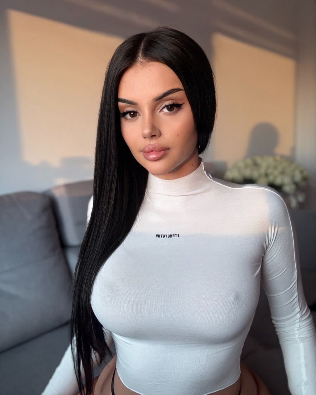 Valentina stands indoors in a white turtleneck with a small logo, near a gray sofa and vase of white flowers.