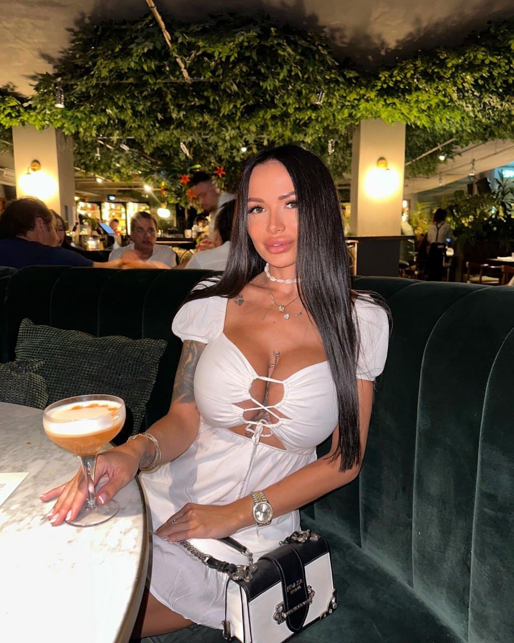 A woman sits in a green velvet booth wearing a white dress with cutouts, holding a stemmed cocktail glass on a marble table.