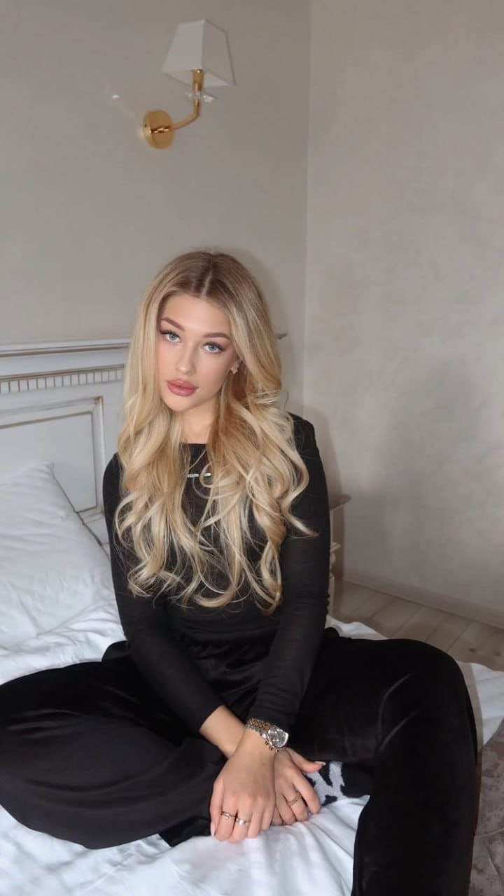 A woman sits cross-legged on a white bed in a black long-sleeved top and pants, with a wall lamp and headboard visible behind her.