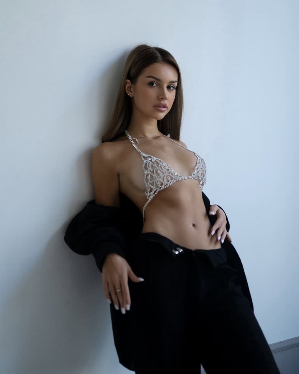 A woman leans against a white wall in a white lace bralette, black trousers, and a black jacket draped off her shoulders.