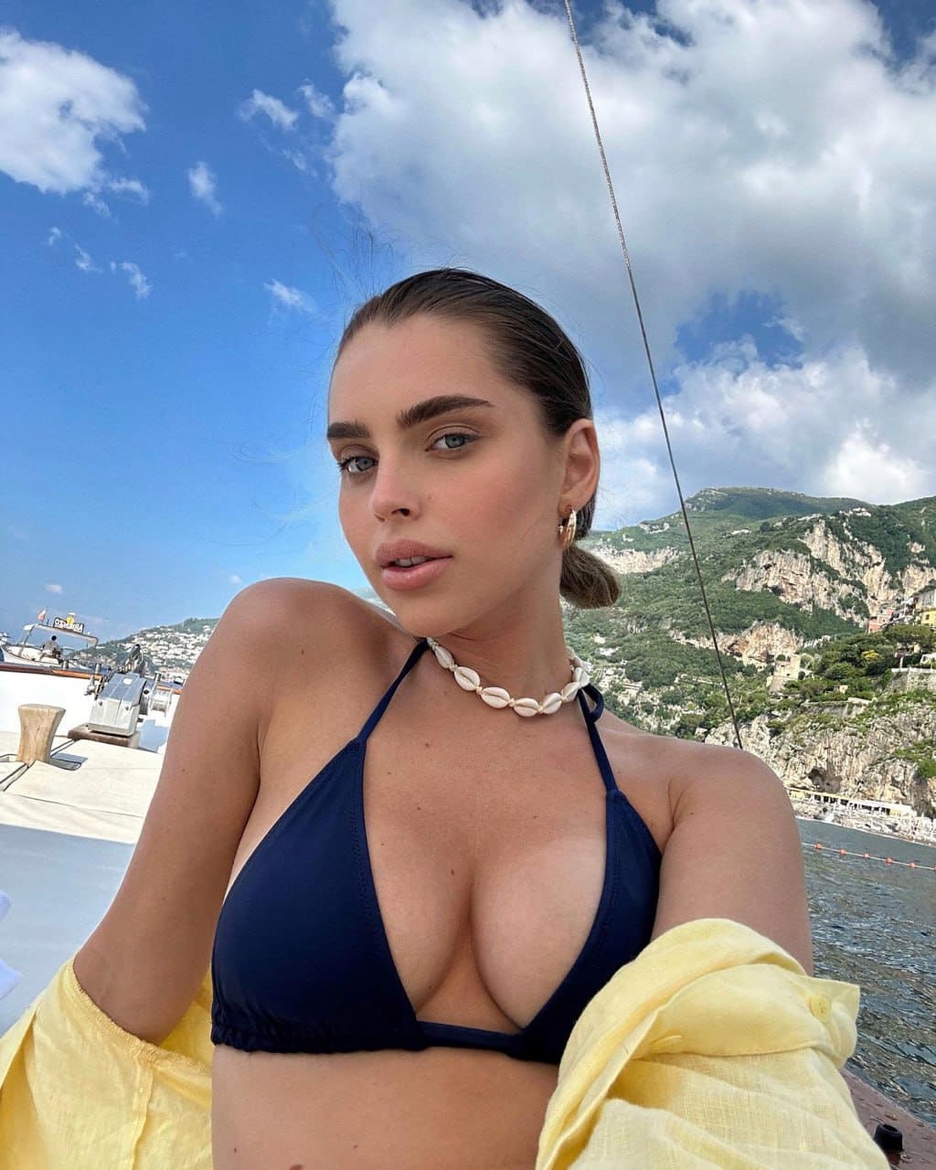 A woman in a navy halter bikini and yellow shirt takes a selfie on a boat with coastal hills behind.