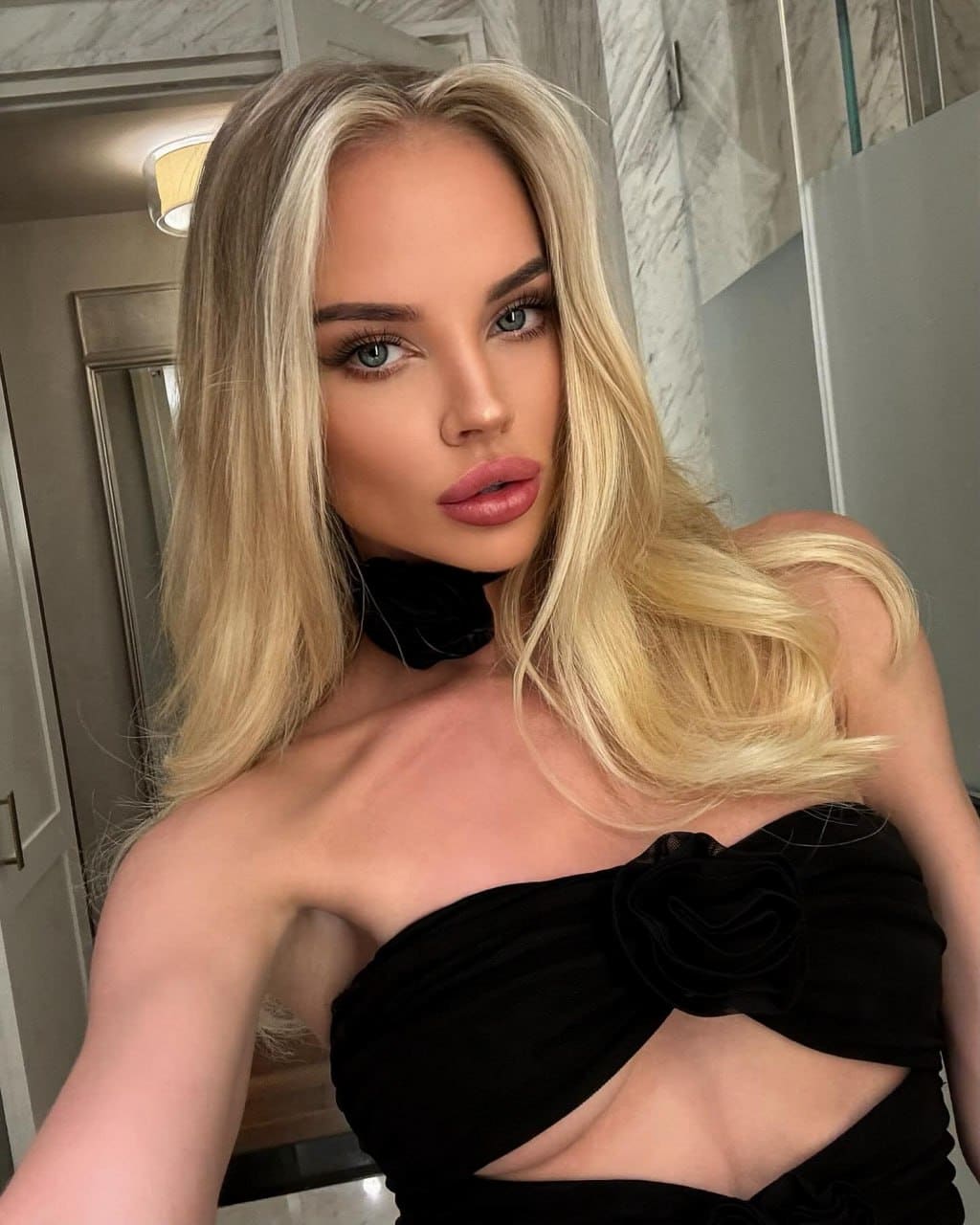 A woman poses in a black strapless top with a midriff cutout and choker, standing in a room with marble walls and a mirror.