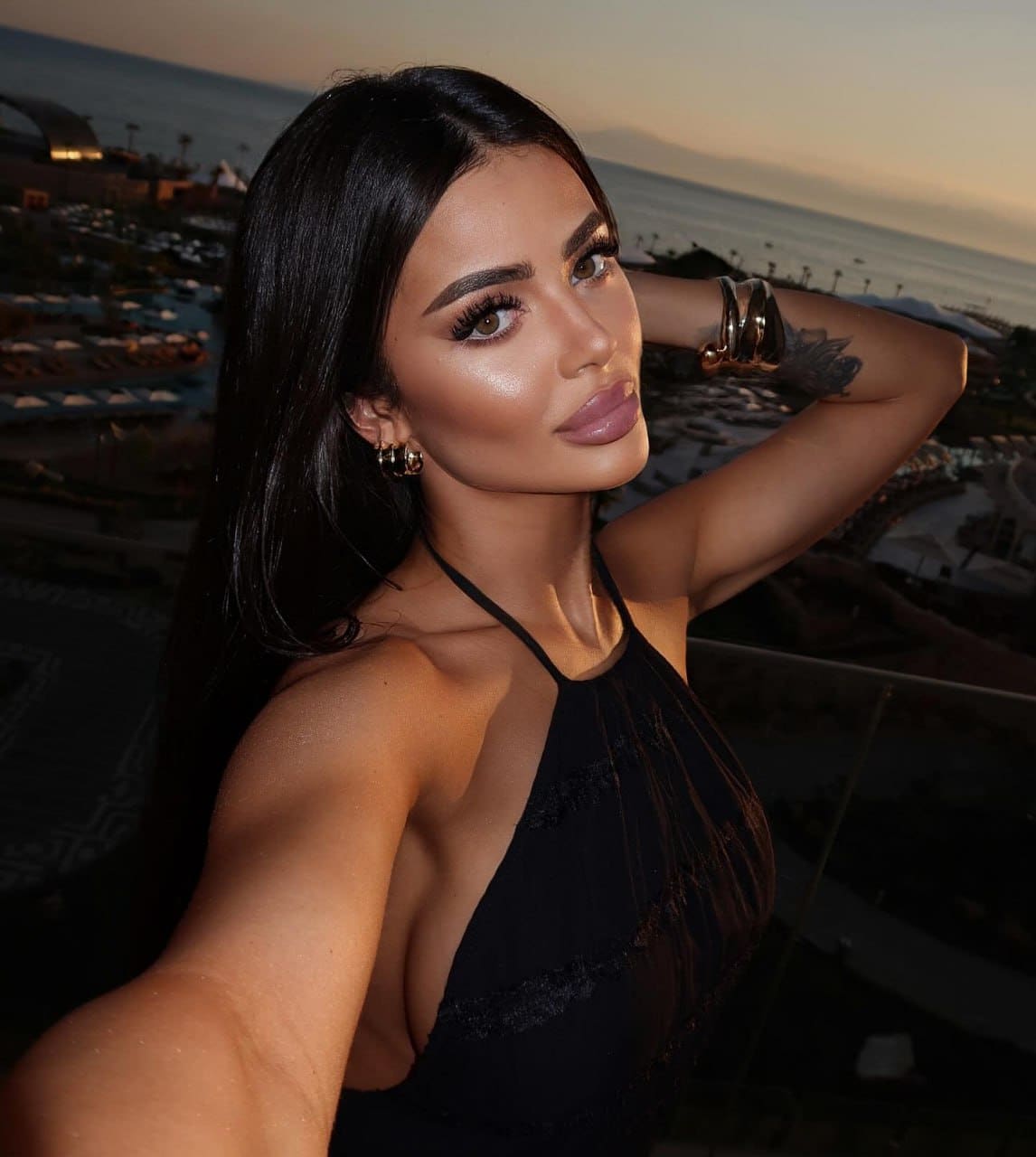 A woman in a black halter top poses with one arm raised against a sunset ocean and resort backdrop.
