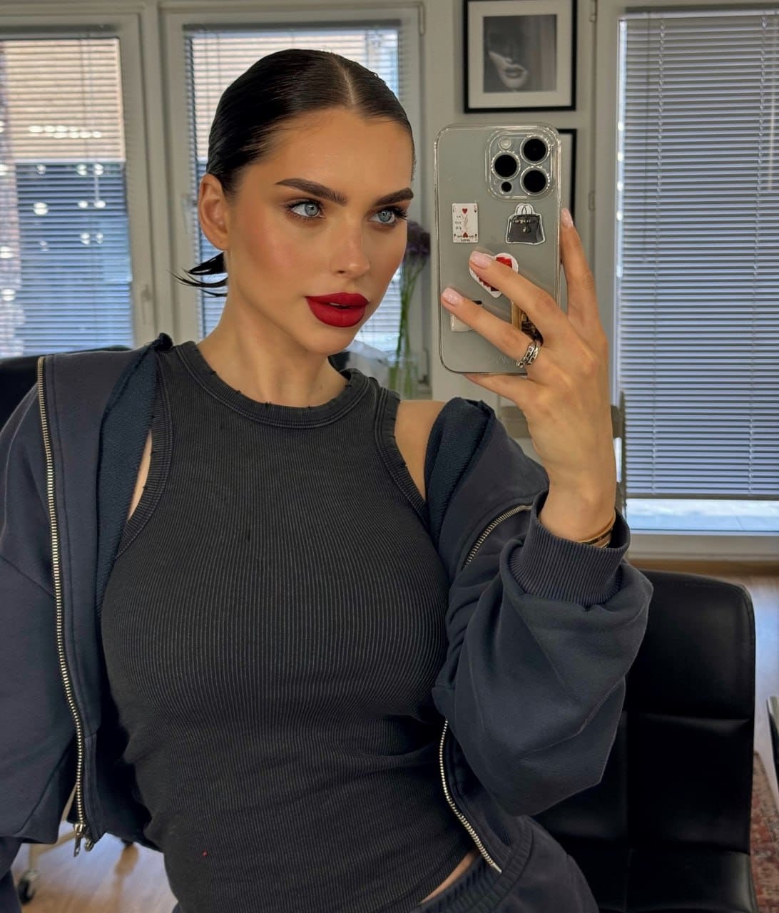 Seated by windows, a woman in a dark ribbed top and open hoodie takes a mirror selfie with a phone.