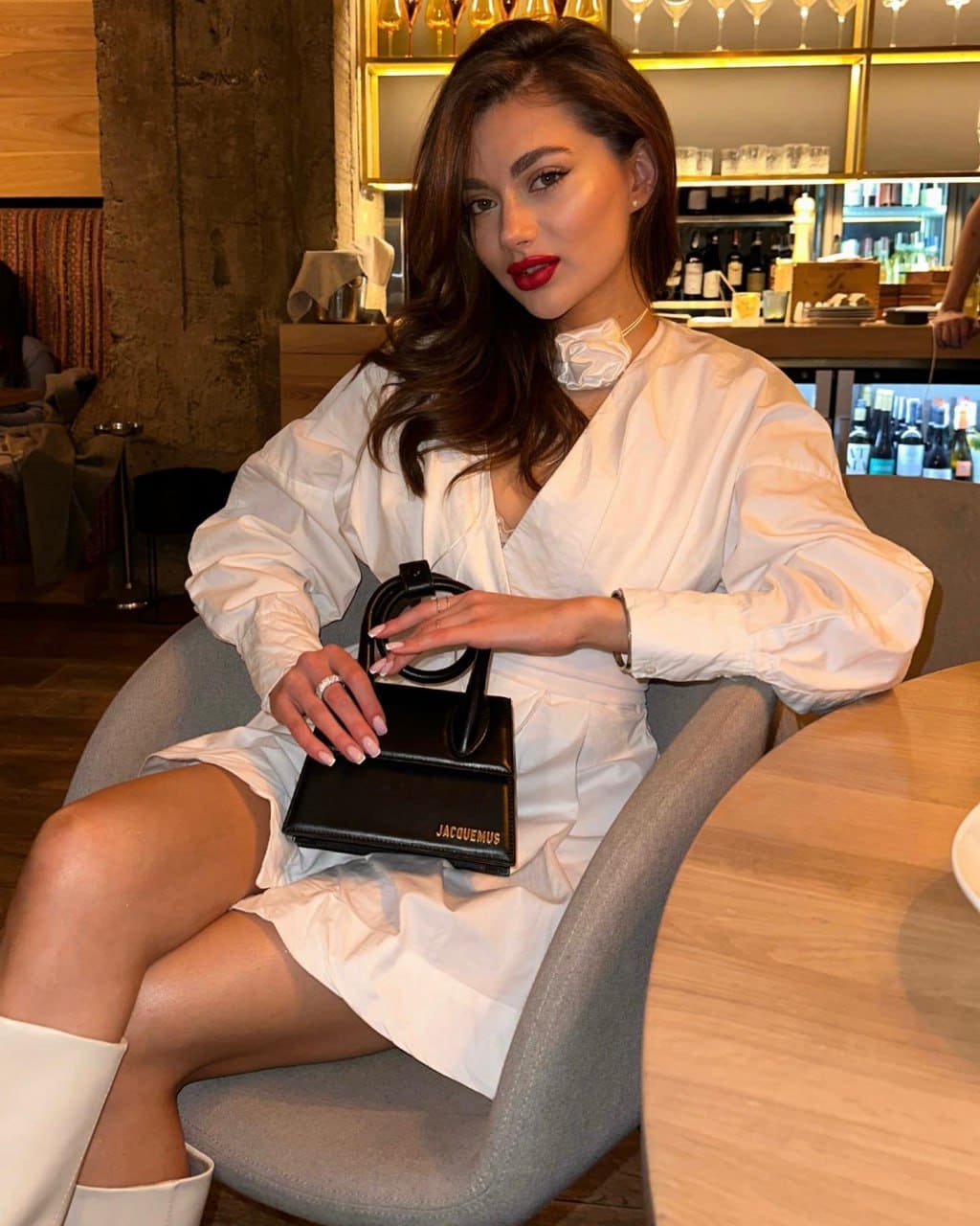 A woman in a white ruffled dress and boots sits in a grey armchair, holding a black handbag at a restaurant table.