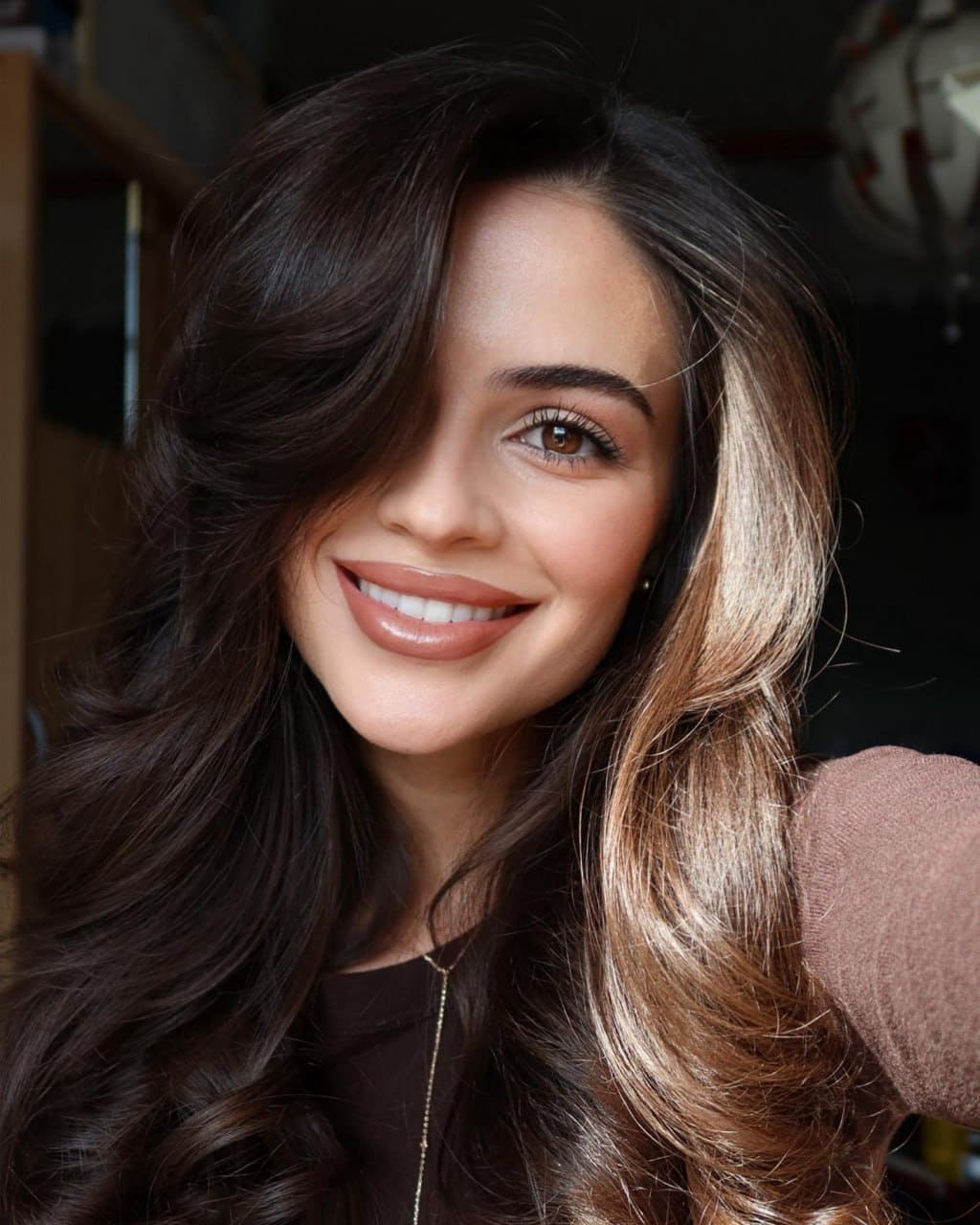 Close-up portrait of a smiling woman with highlighted wavy hair, wearing a dark top and gold necklace indoors.