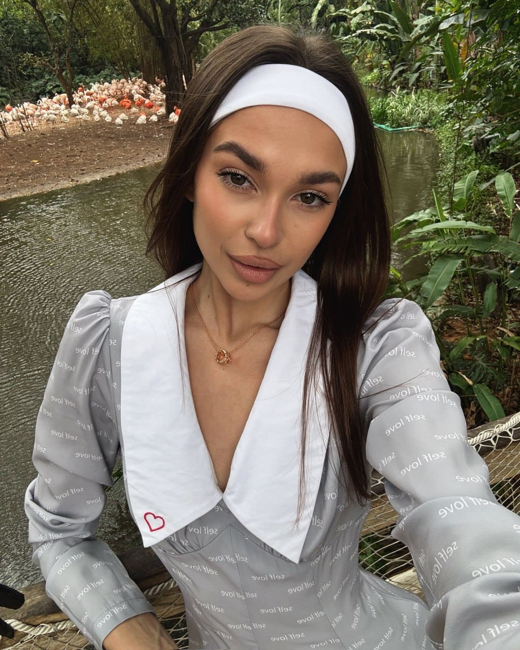 A woman in a grey dress with a white collar takes a selfie near a pond, with chickens visible in the background.