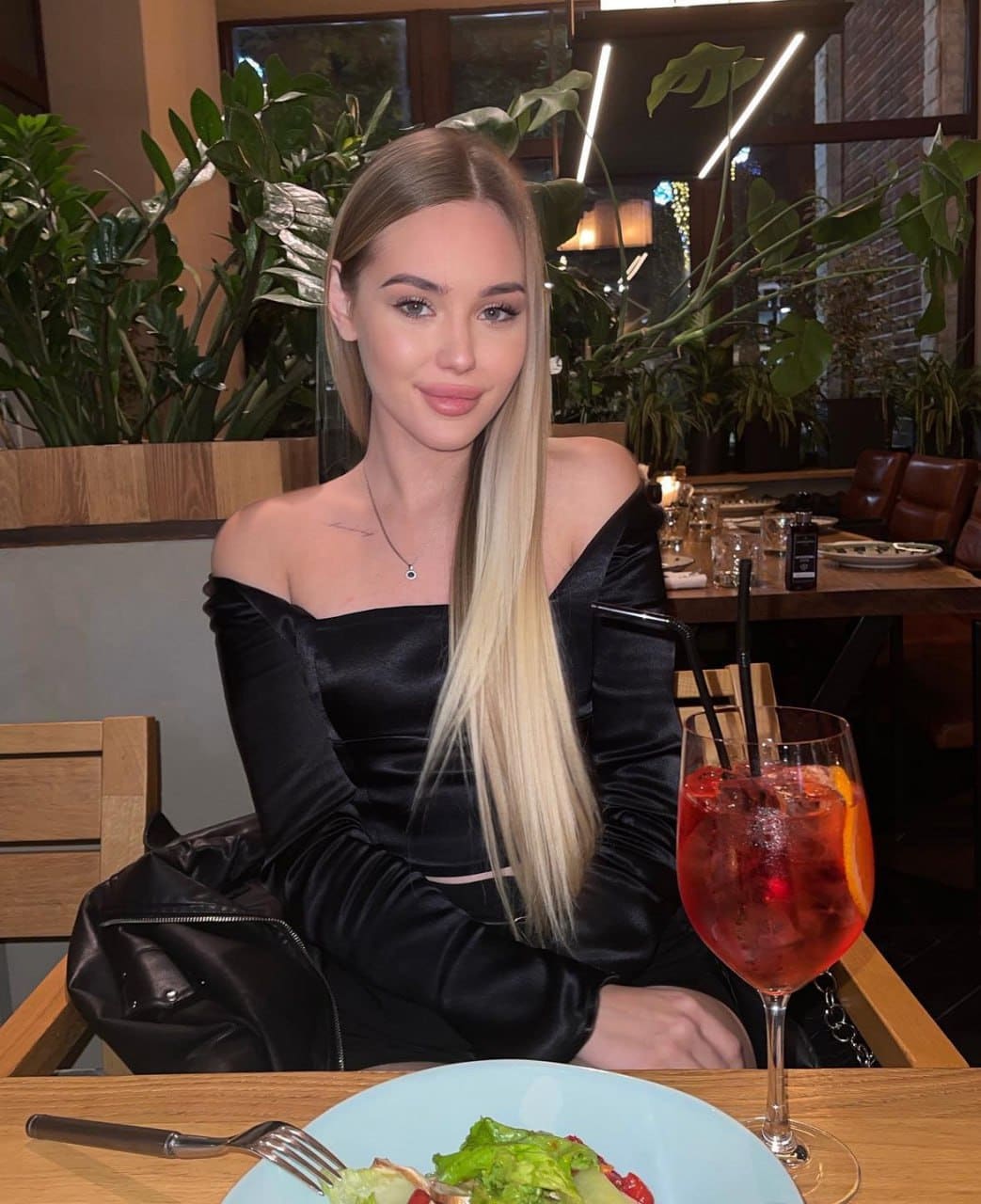 Woman in a black off-the-shoulder satin top sits at a plant-filled restaurant table with red drink and salad.