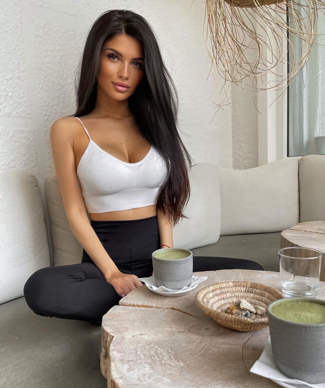 A woman sits cross-legged on a light sofa in a white crop top and black leggings, with matcha, water, and a basket of stones on a table before her.