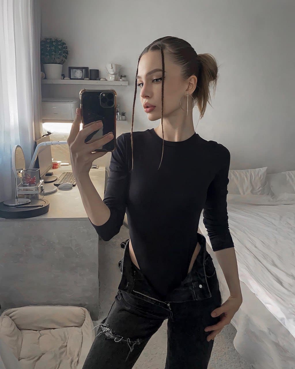 Mirror selfie in a bedroom with a high ponytail, wearing a black long-sleeved top with side cutouts and distressed jeans near a desk.