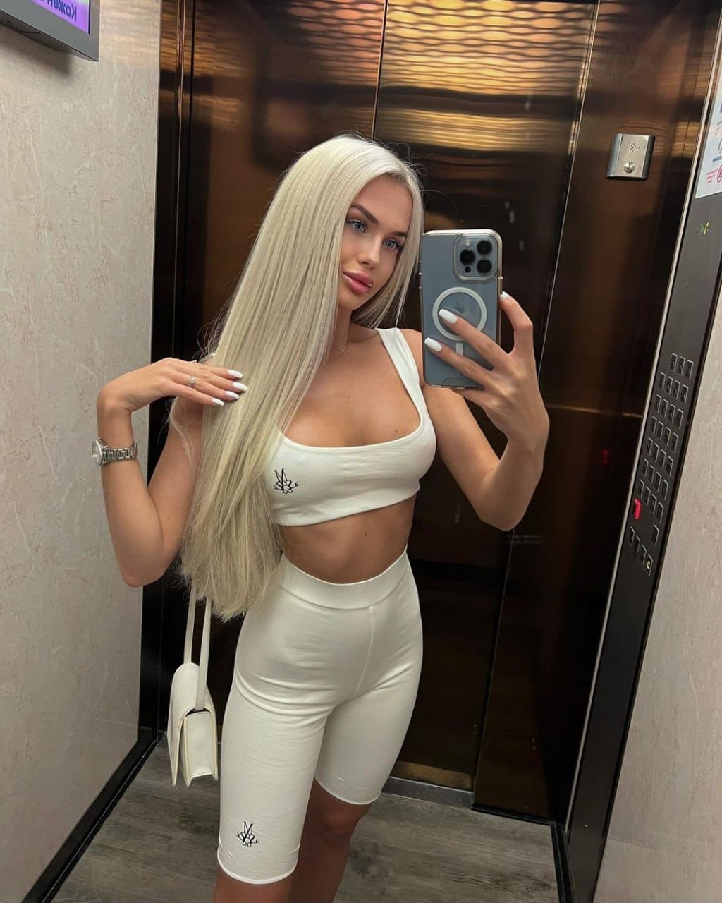 A blonde woman in a white crop top and shorts takes an elevator mirror selfie, holding a phone and white bag.