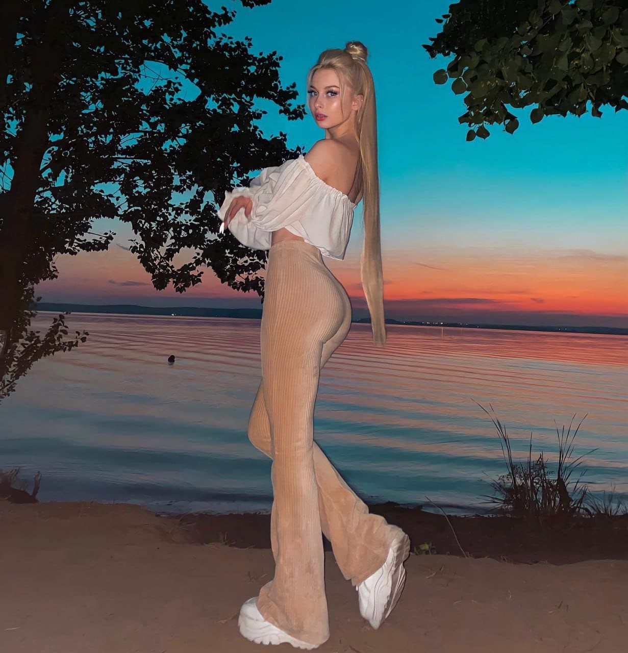Profile view of a blonde woman in a white crop top and beige flared pants on a sandy shore at sunset, with water behind.