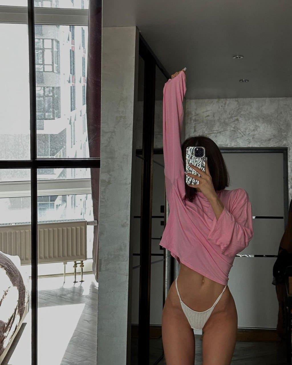 A woman stands by large windows in a cropped pink top and white bottoms, holding a phone for a mirror selfie with arm raised.