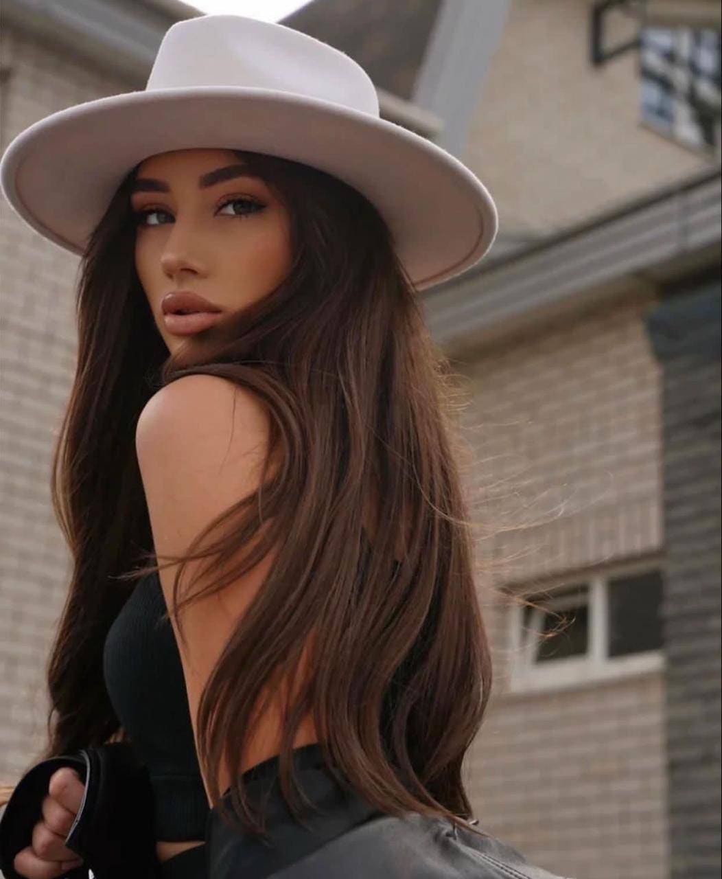 A woman in a wide-brimmed white hat and black sleeveless top looks over her shoulder while standing outdoors before a brick building.