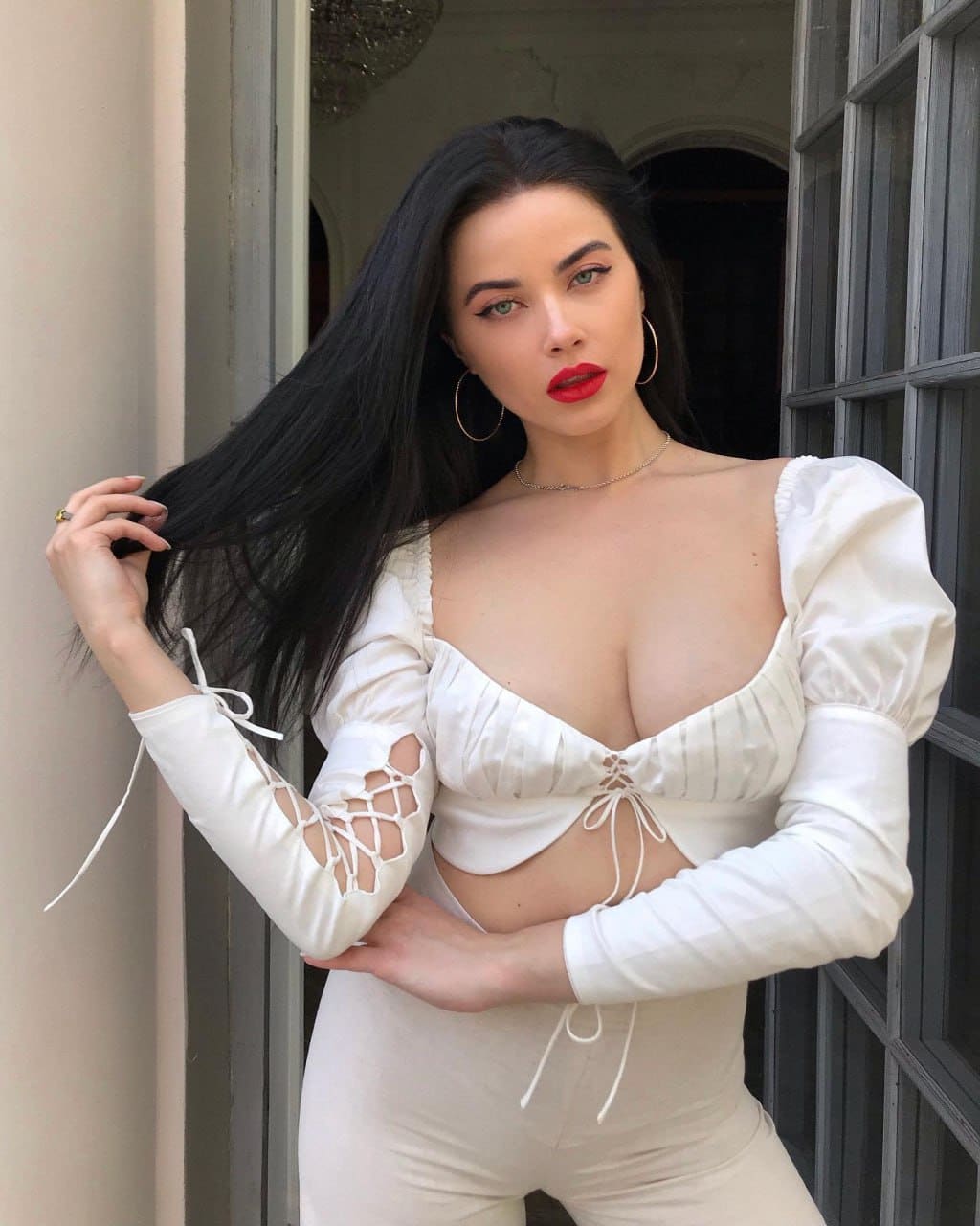 A woman stands in a doorway wearing a white two-piece outfit with puffed sleeves and high-waisted bottoms, holding a strand of her dark hair.