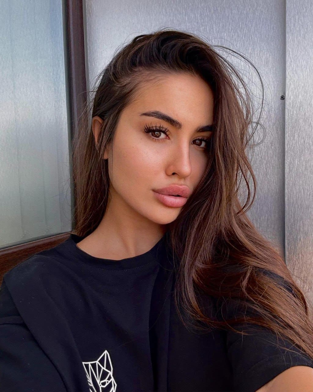 Isabella with long wavy dark hair wears a black t-shirt with a white geometric drawing, standing before a metal wall and window frame.