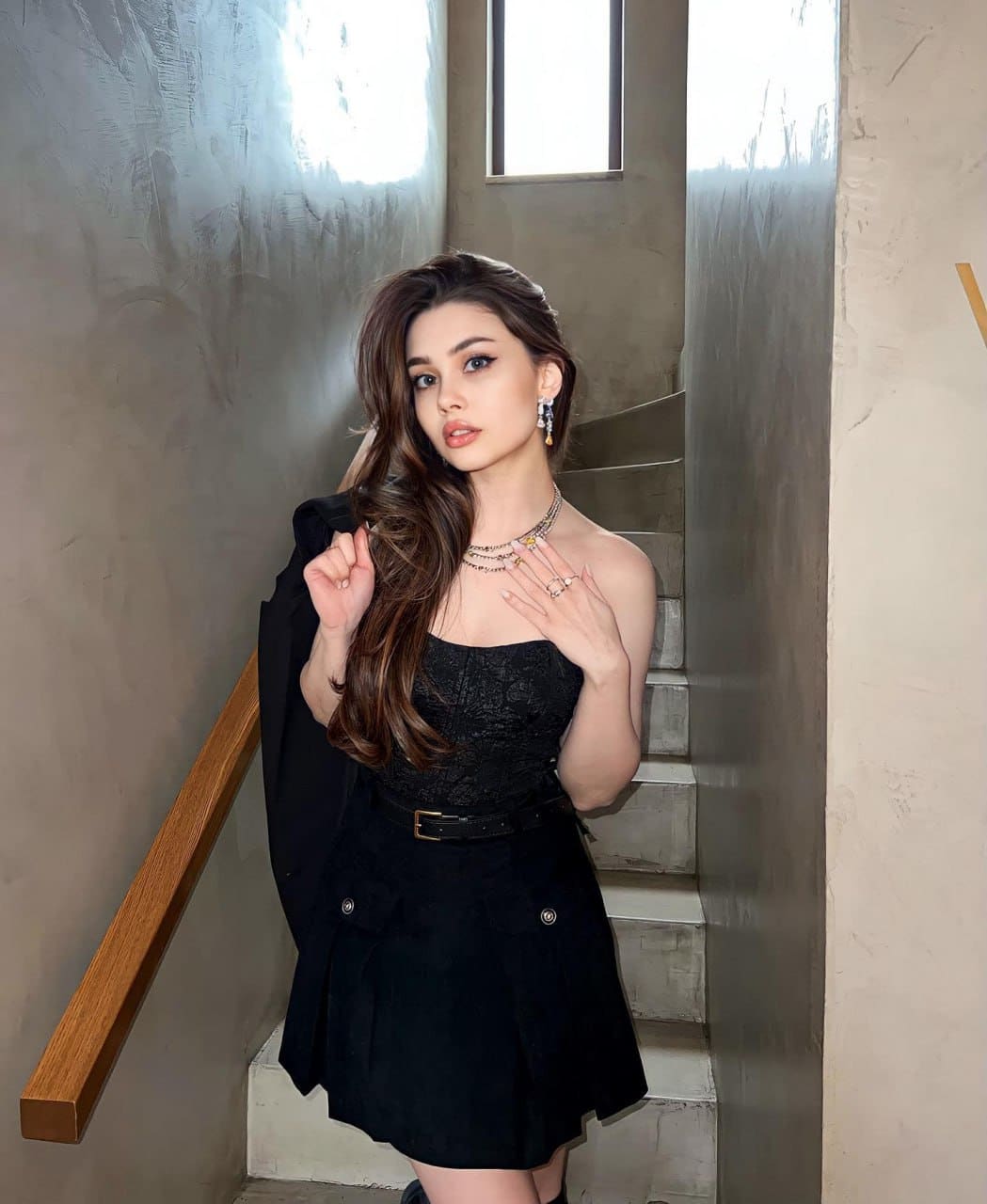 Mia stands on concrete stairs in a black lace top, mini skirt, and jacket draped over one shoulder, wearing layered necklaces.