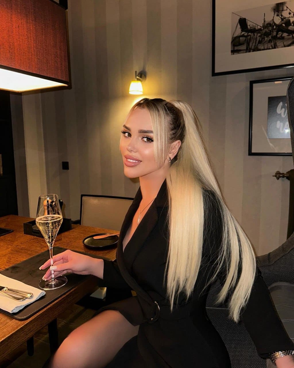 A woman with a high blonde ponytail sits at a wooden table in dim light, wearing a black blazer dress and holding a glass of sparkling wine.