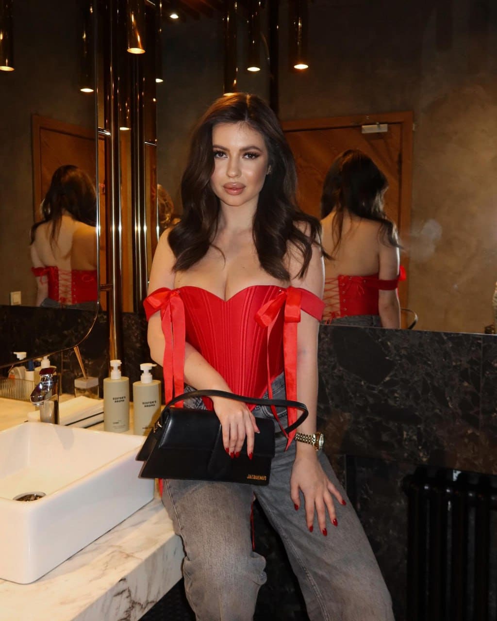 A woman in a red off-the-shoulder pleated top and grey jeans leans against a marble bathroom counter, holding a black handbag.
