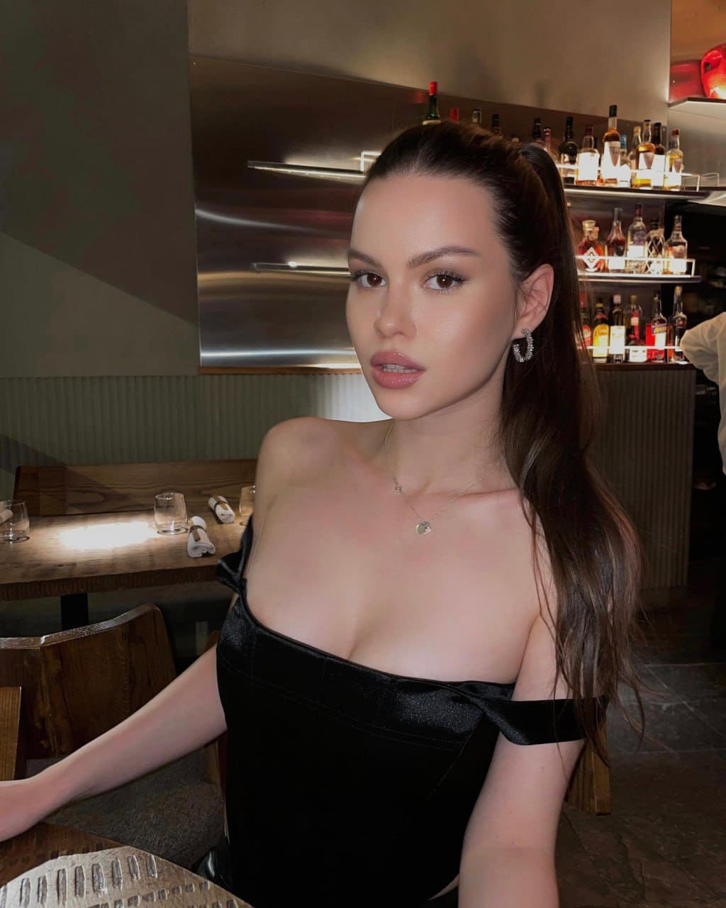 A woman with a high ponytail sits at a bar table in a black off-the-shoulder top, with shelves of liquor bottles visible in the background.