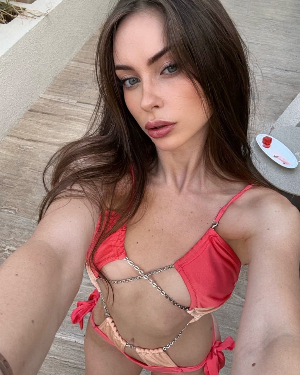 A woman takes a selfie on a wooden deck in a red and beige string bikini, standing beside a table with food.