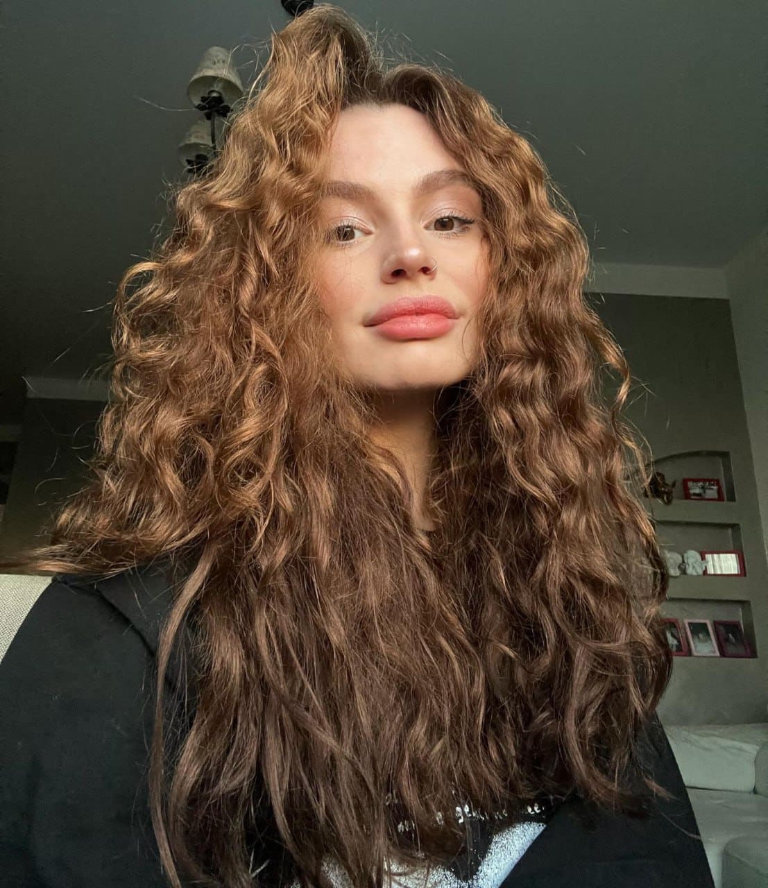 A person with voluminous curly hair wears a dark jacket over a light top, posing indoors near shelves and a ceiling light.