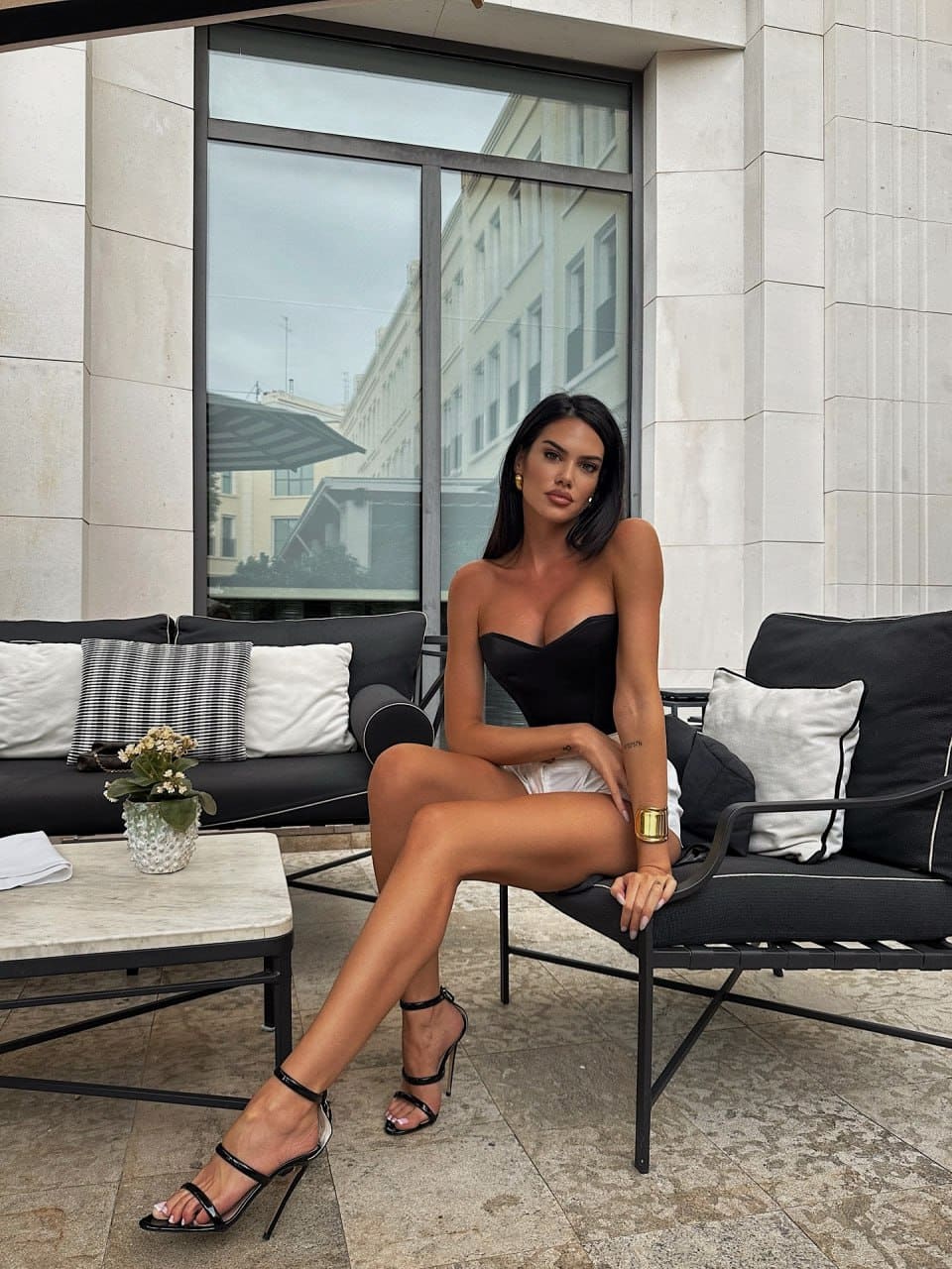 A woman sits on a black lounge chair in a black top and white shorts, with a marble table behind her.