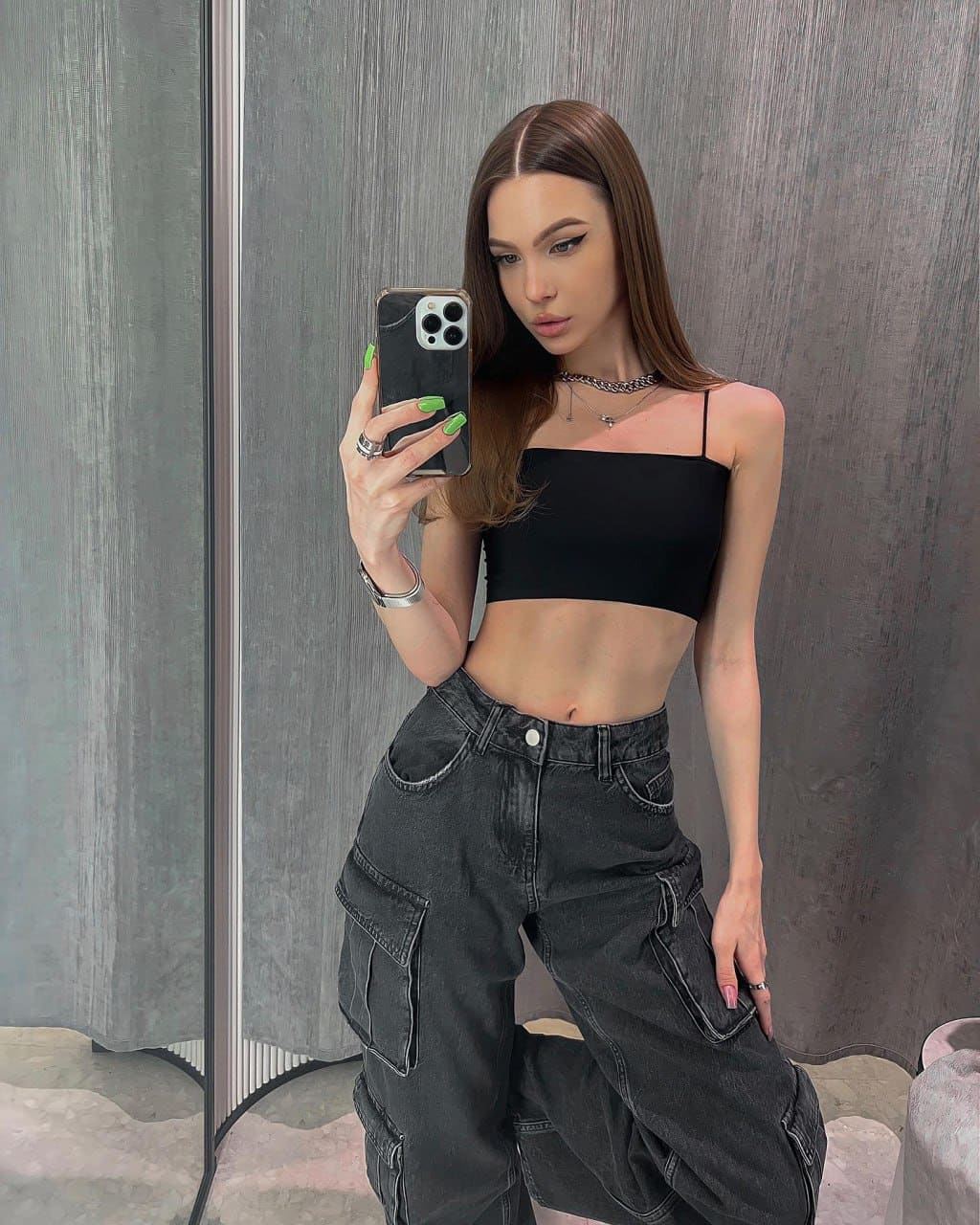 Mirror selfie in a gray room, wearing a black spaghetti-strap crop top and dark cargo jeans, holding a phone with green nails.