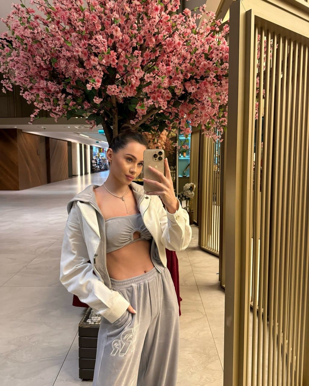 Mirror selfie of a woman in a grey crop top, sweatpants, and open white jacket, standing in a corridor before a pink flowering tree.