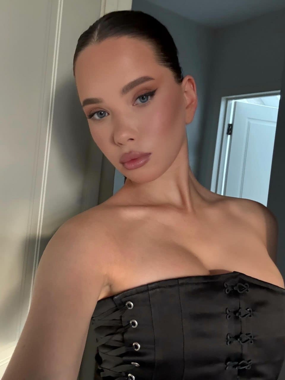 A woman with sleek dark hair poses indoors near a white door frame wearing a black strapless corset top with lace-up detailing.