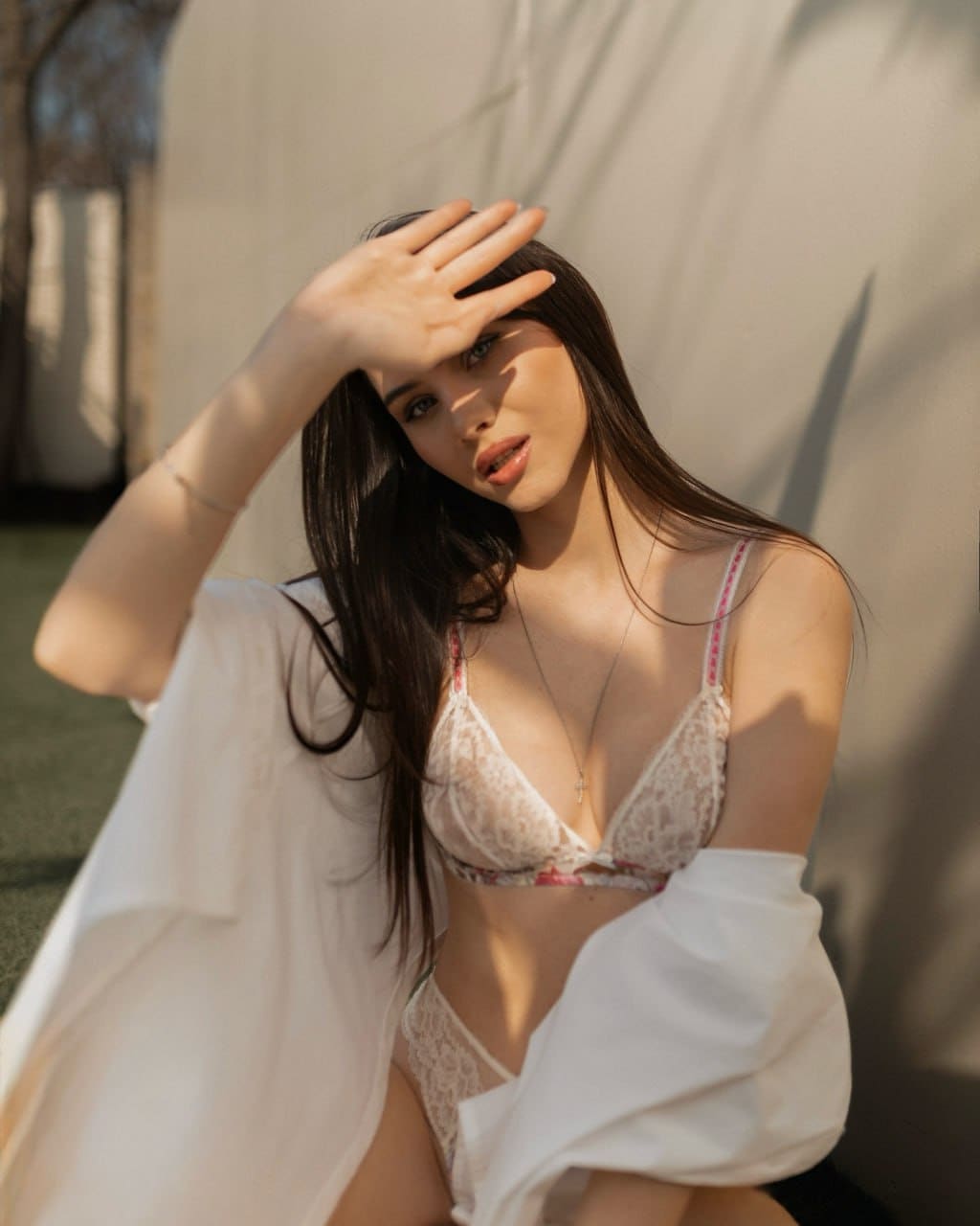 She sits outdoors against a light wall in white lace lingerie and a draped shirt, raising one hand to shade her eyes from the sun.