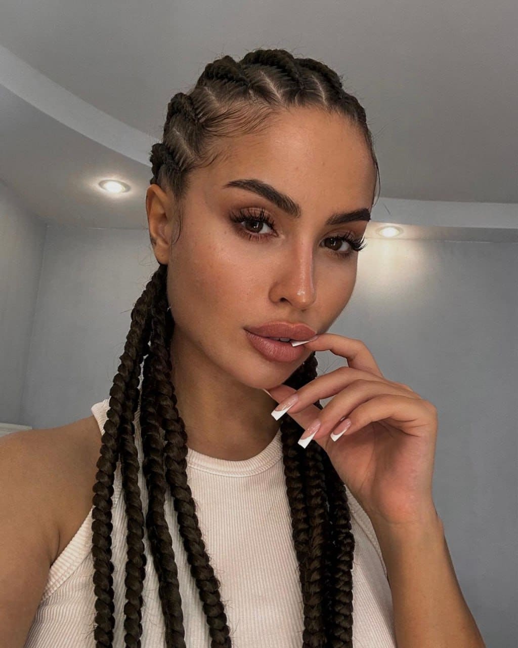 A woman with cornrowed hair wears a white ribbed tank top, posing with a hand near her mouth in a room with ceiling lights.