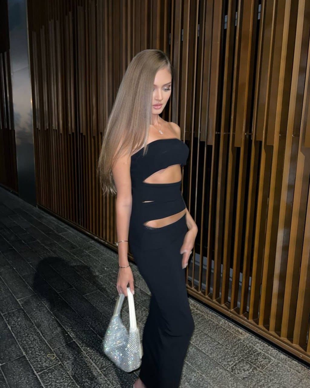 Woman in profile wears a black cut-out top and trousers, holding a silver beaded bag against a vertical wooden slat wall outdoors.