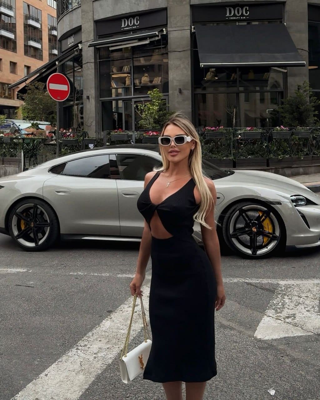A blonde woman stands on a city street before a silver sports car, wearing a black cut-out midi dress, white sunglasses, and holding a white handbag.