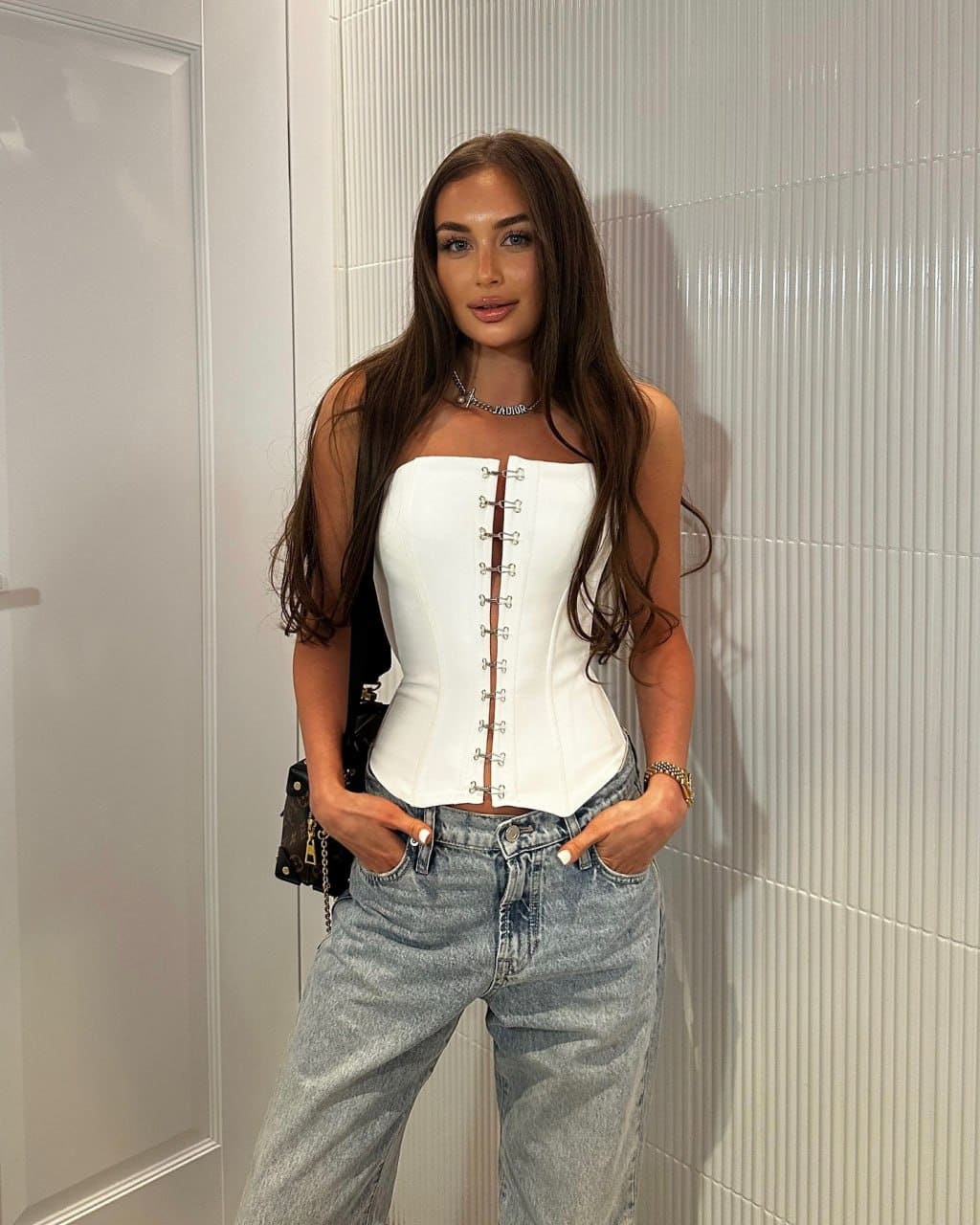 Violetta stands against a white ribbed wall in a white strapless corset top with front lacing and light-wash jeans, hands in pockets.