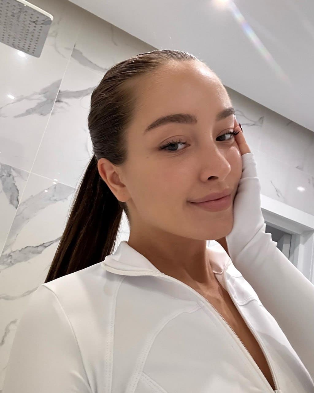 A woman in a white jacket and gloves rests her hand on her cheek in a marble bathroom with a shower head.