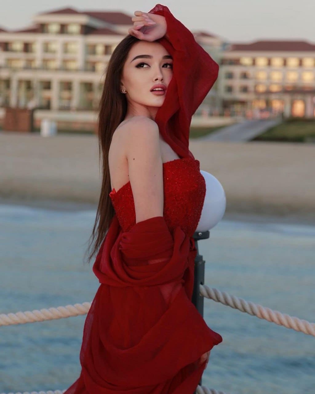 A woman stands in profile on a waterfront promenade in a red sequined strapless dress with draped fabric, hand raised to her head.