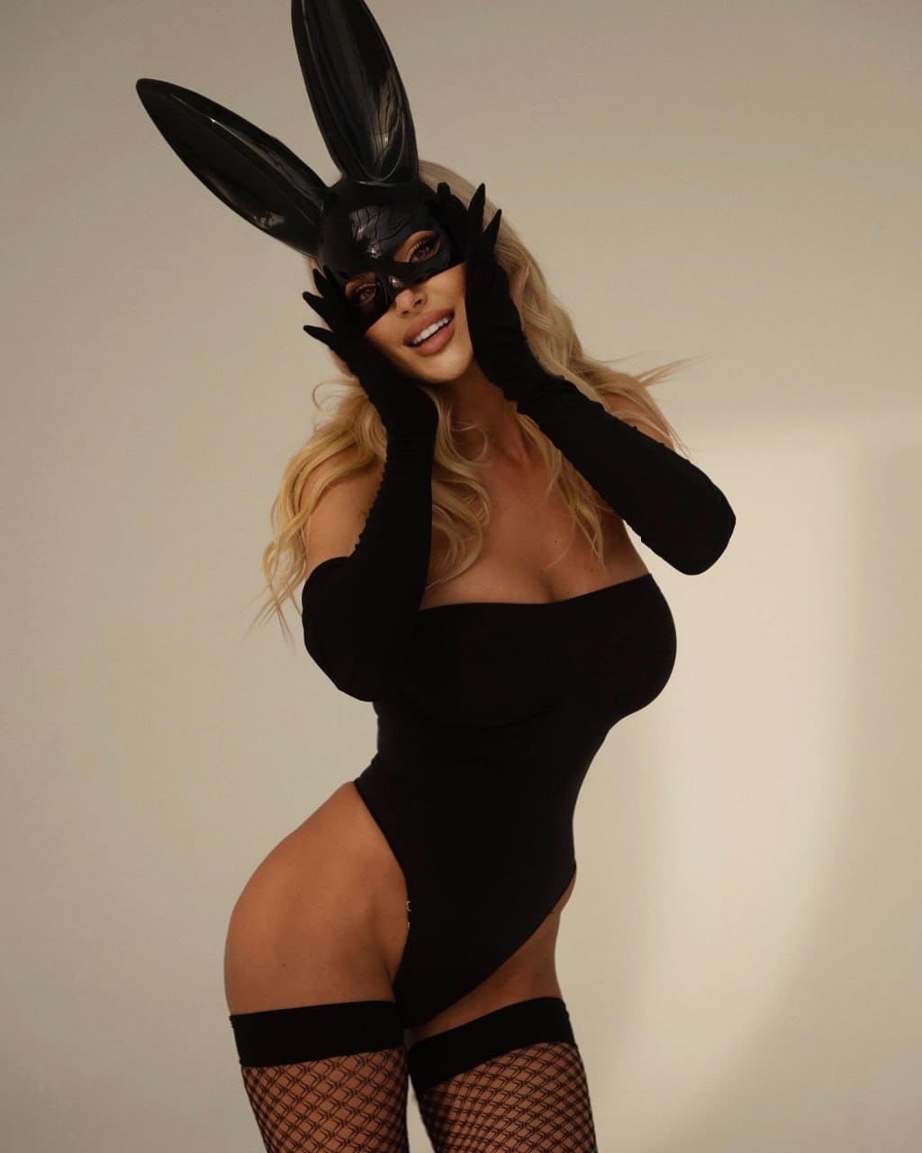 A blonde woman in a black bodysuit, fishnets, gloves, and rabbit ears poses against a plain background.