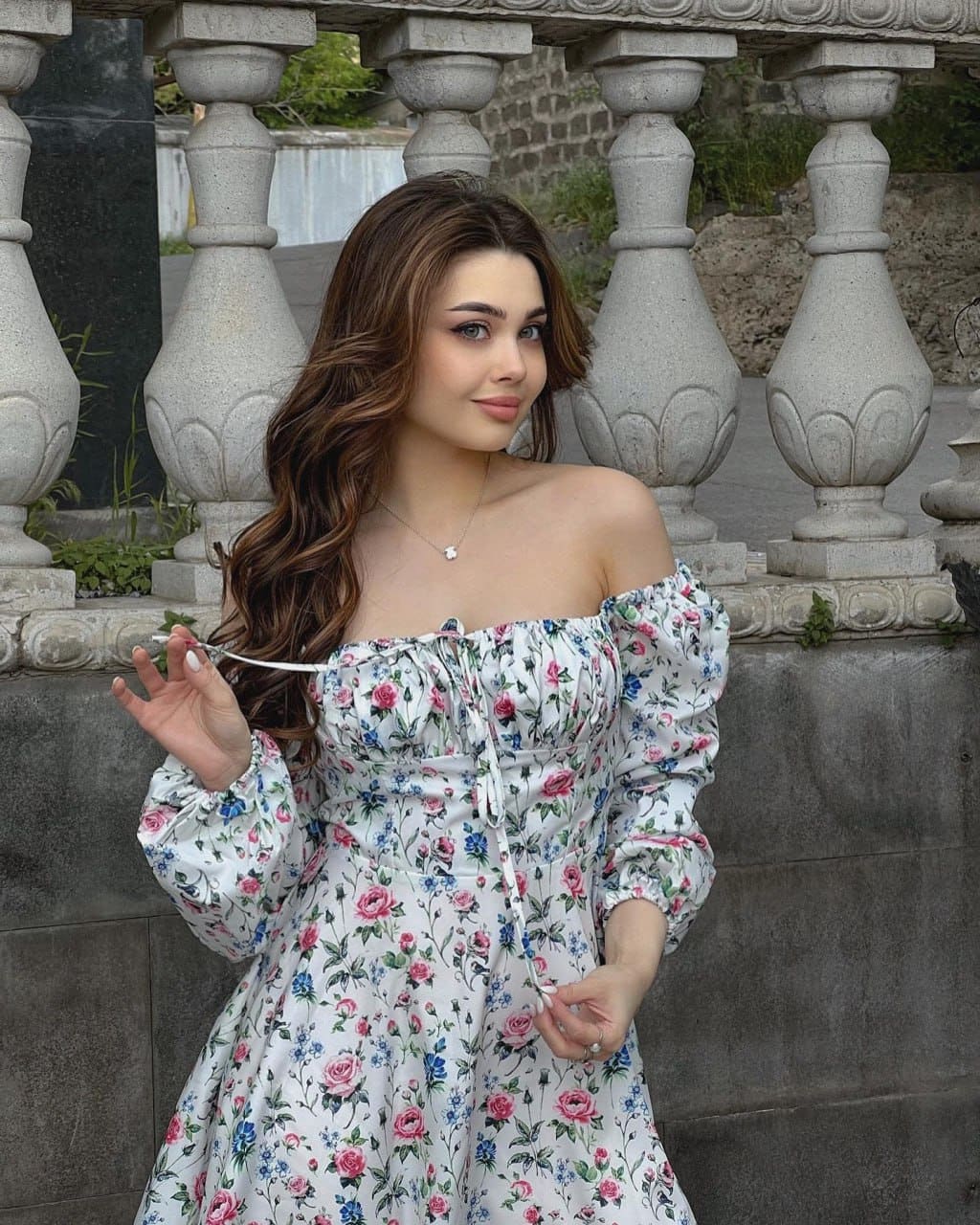A woman in a white off-the-shoulder floral dress stands outdoors by a stone balustrade, holding a thin white strap.