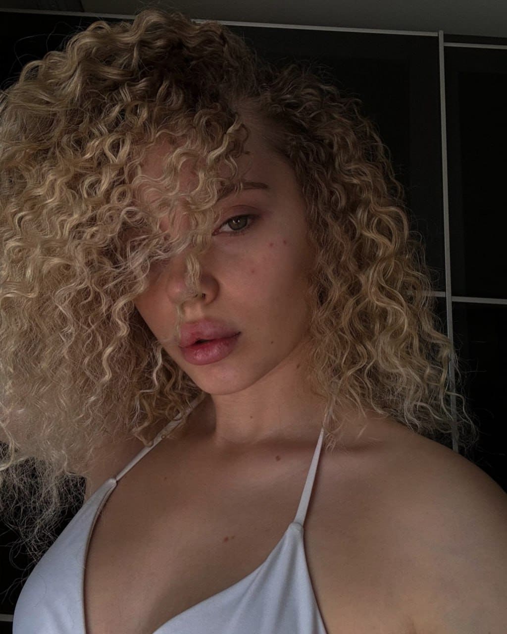 Close-up of a person with voluminous curly blonde hair partially covering their face, wearing a white halter-neck top against a dark paneled background.