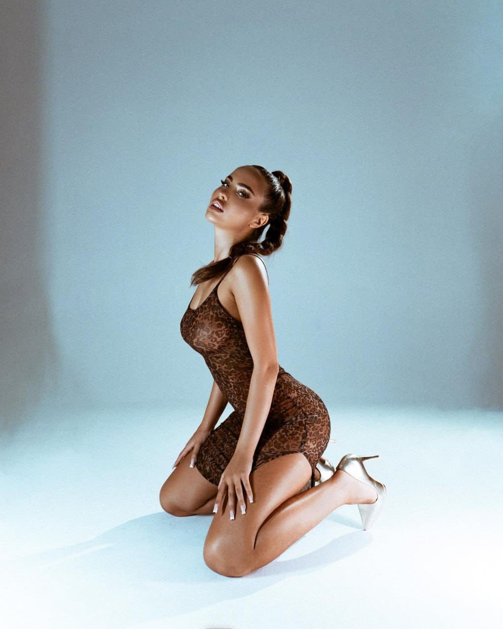 A woman in a leopard-print dress and gold heels kneels on a light floor against a blue-gray backdrop, head tilted back.