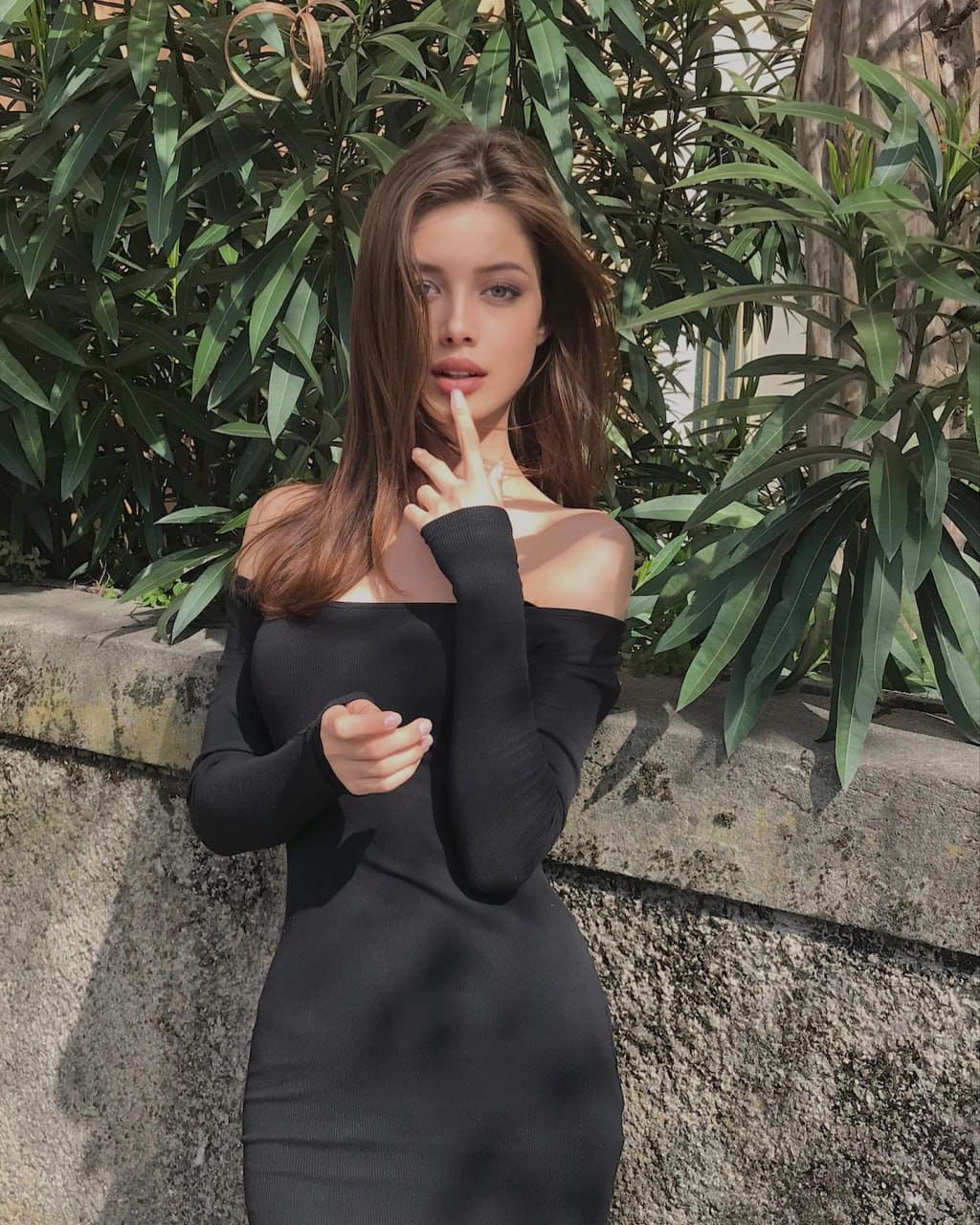 Woman in a black off-the-shoulder dress stands before a stone wall and greenery, holding a finger to her lips.