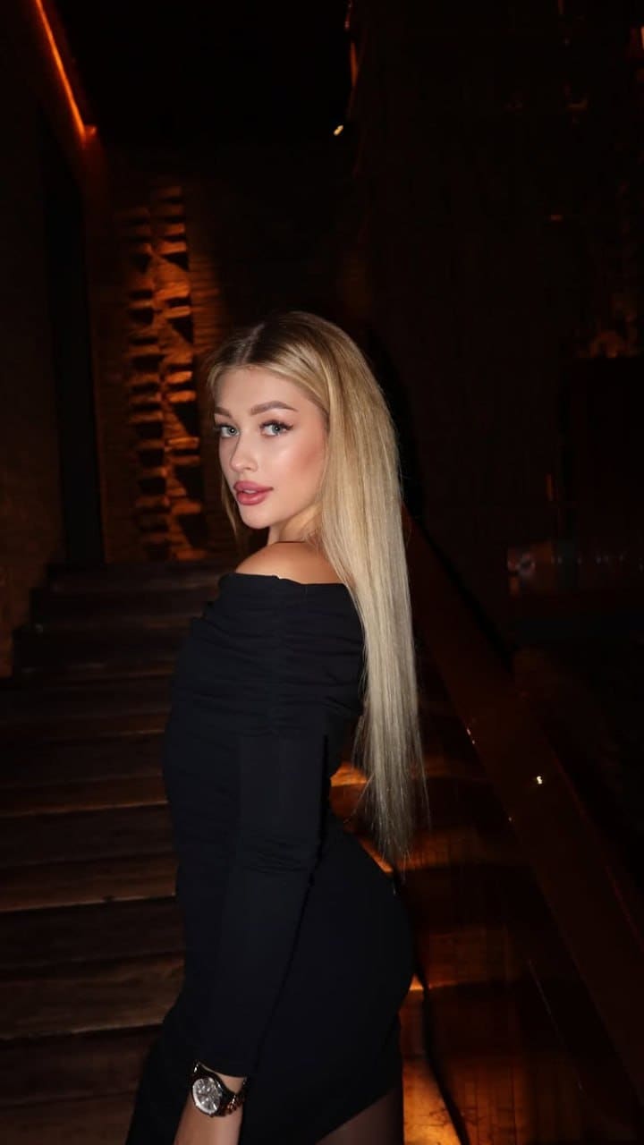 In profile on a dim wooden staircase, a woman in a black off-the-shoulder dress looks back over her shoulder, wearing a wristwatch.