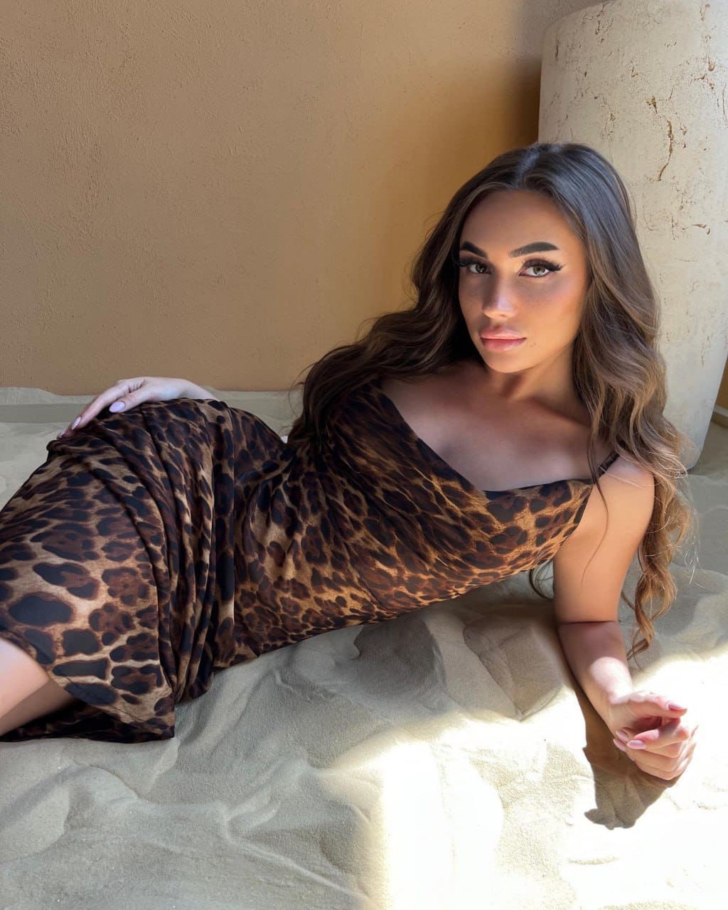 A woman lies on her side on sand, propped on one arm in a leopard-print dress, against a tan wall and stone pillar.