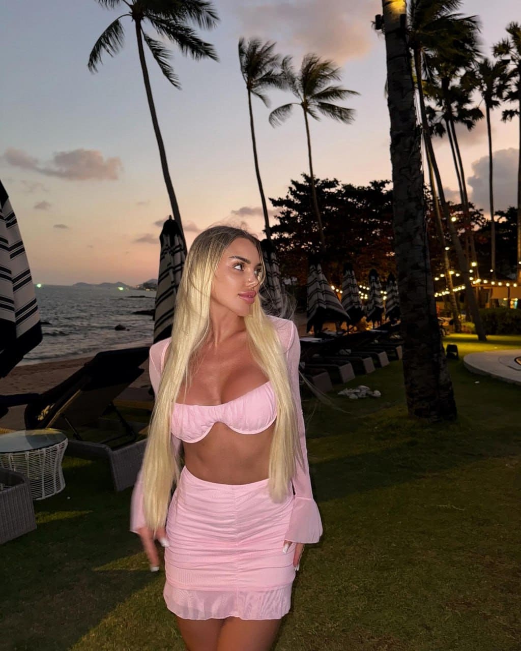 A blonde woman stands on grass near a beach at dusk in a pink bandeau top, short skirt, and sheer cardigan, with palm trees in the background.