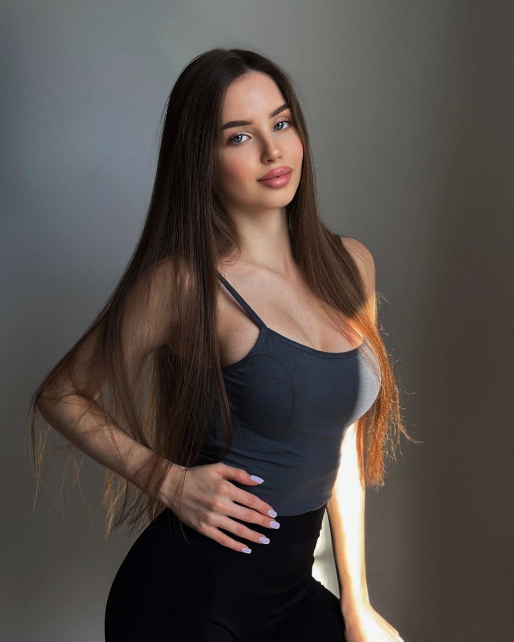 A woman stands in a studio against a gray backdrop, wearing a dark gray tank top and black high-waisted bottoms with a hand on her hip.