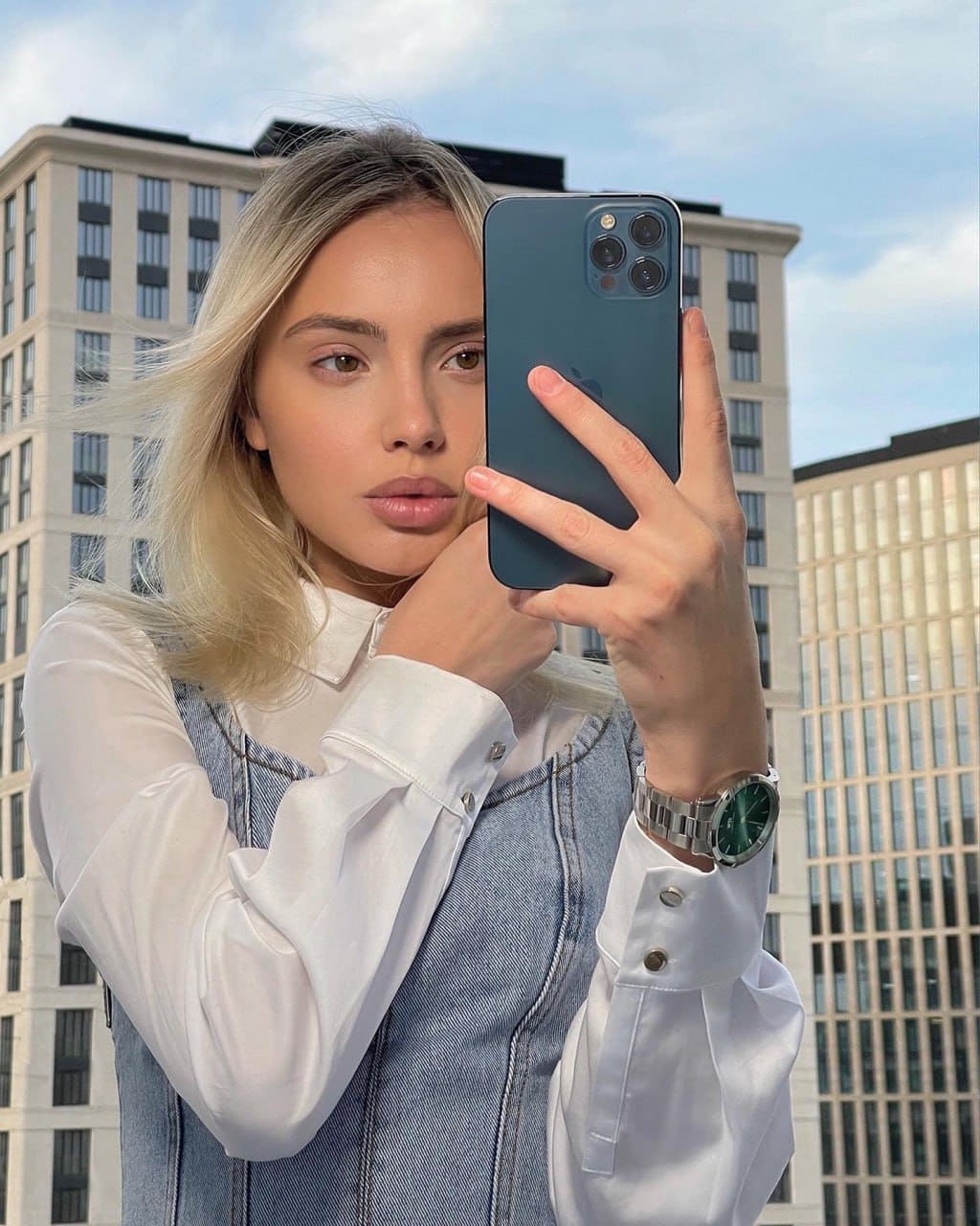 A mirror selfie shows a blonde in a white shirt under a denim overall dress, holding a phone, with city buildings and sky in the background.
