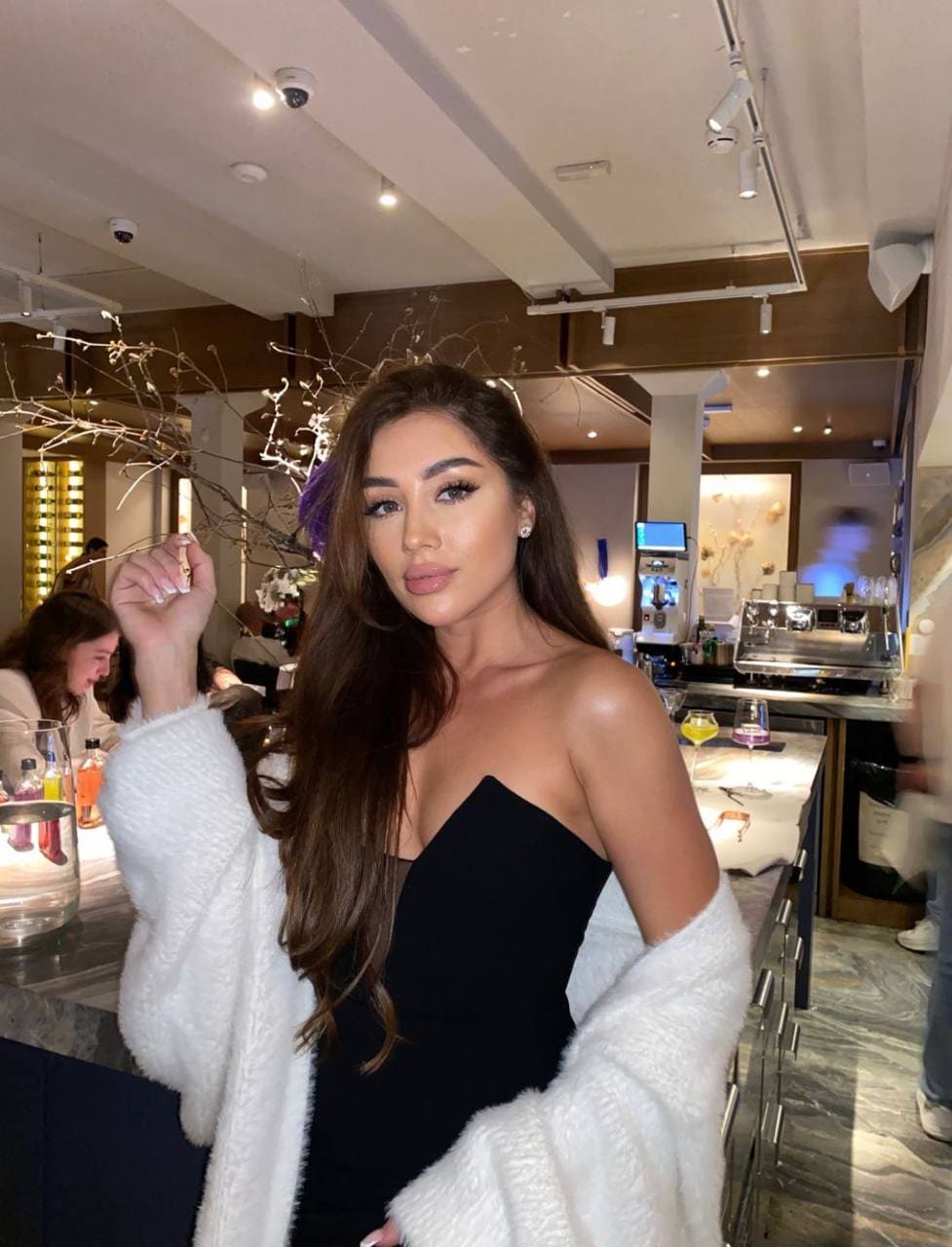Elianora stands at a bar in a black strapless top and white cardigan, hand near head, with track lighting and a coffee machine behind.