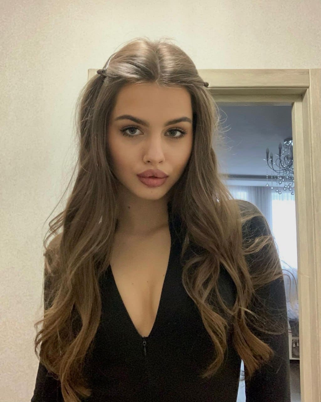 A woman with light-brown wavy hair wears a black long-sleeved V-neck top, standing indoors near a doorway with a chandelier visible.