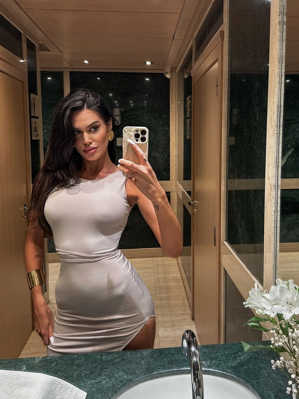 A woman stands by a green marble sink in a light sleeveless dress with a slit, wearing gold jewelry.